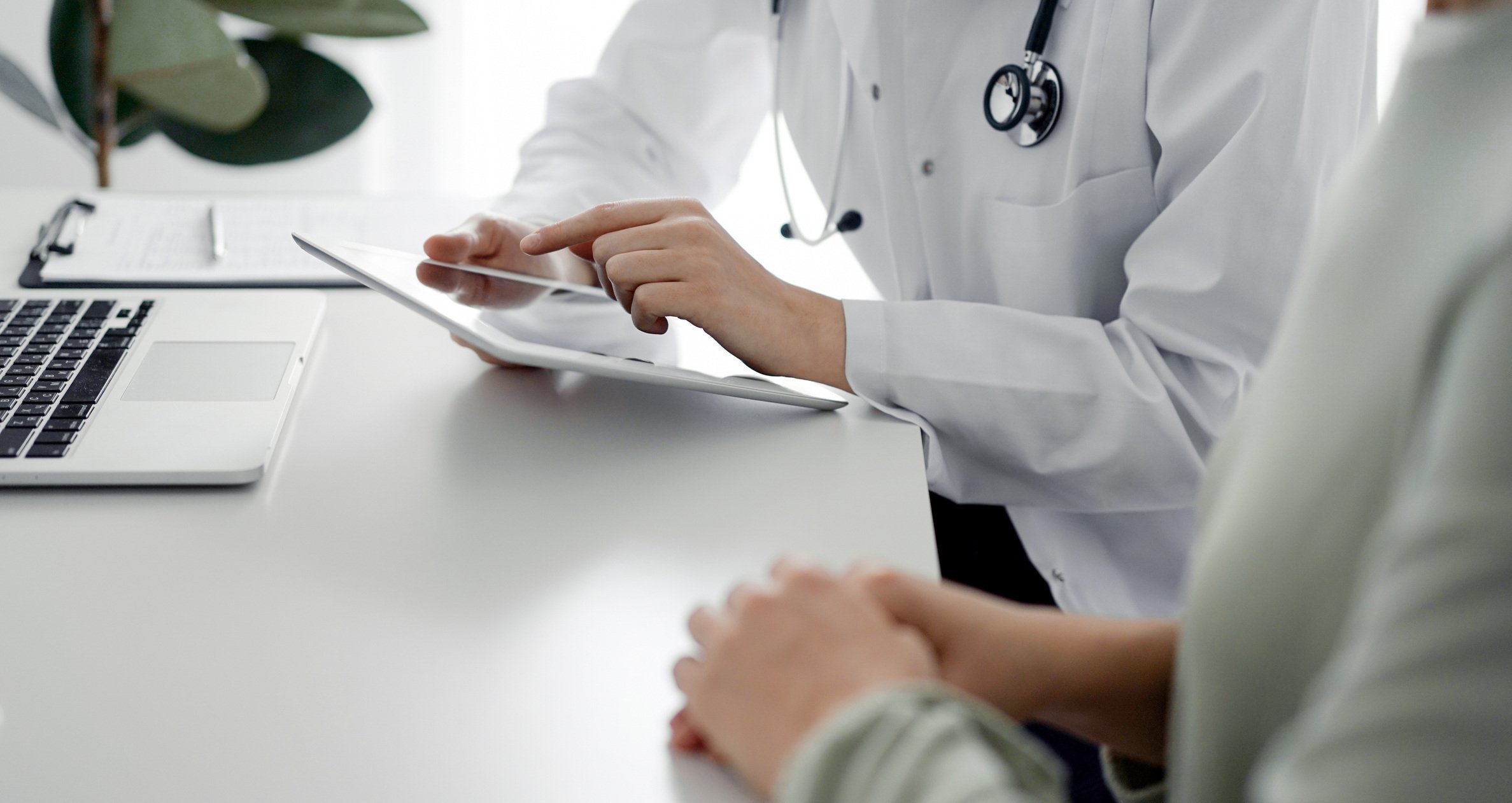 Doctor and patient sitting at the desk in clinic office. The focus is on female physician's hands using tablet computer, close up. Medicine concept.