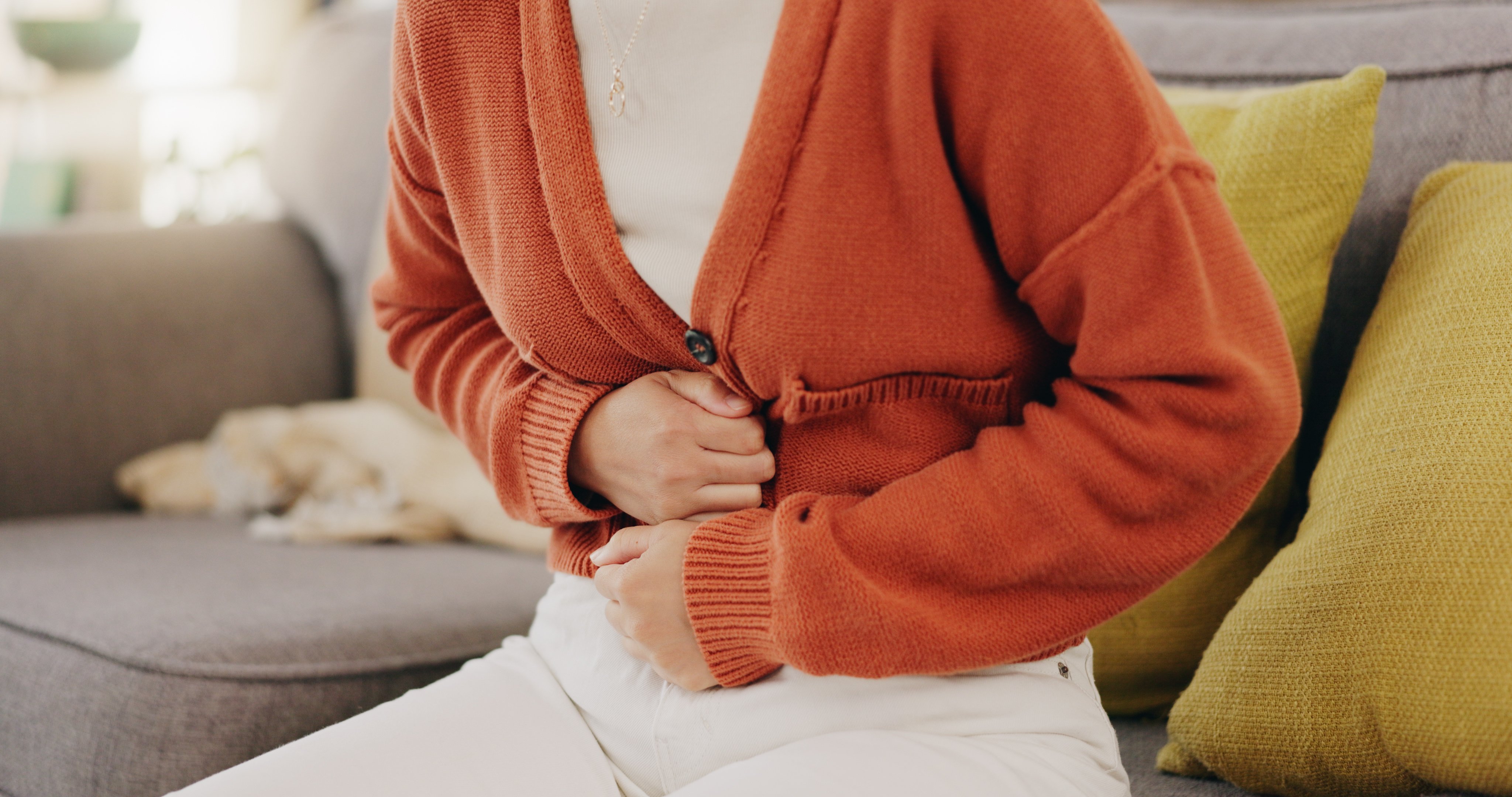 Stomach ache, cramps and hands of woman with abdomen pain due to constipation, menstruation or ibs issue. Sick, home and person suffering and holding belly in a house lounge, couch and living room