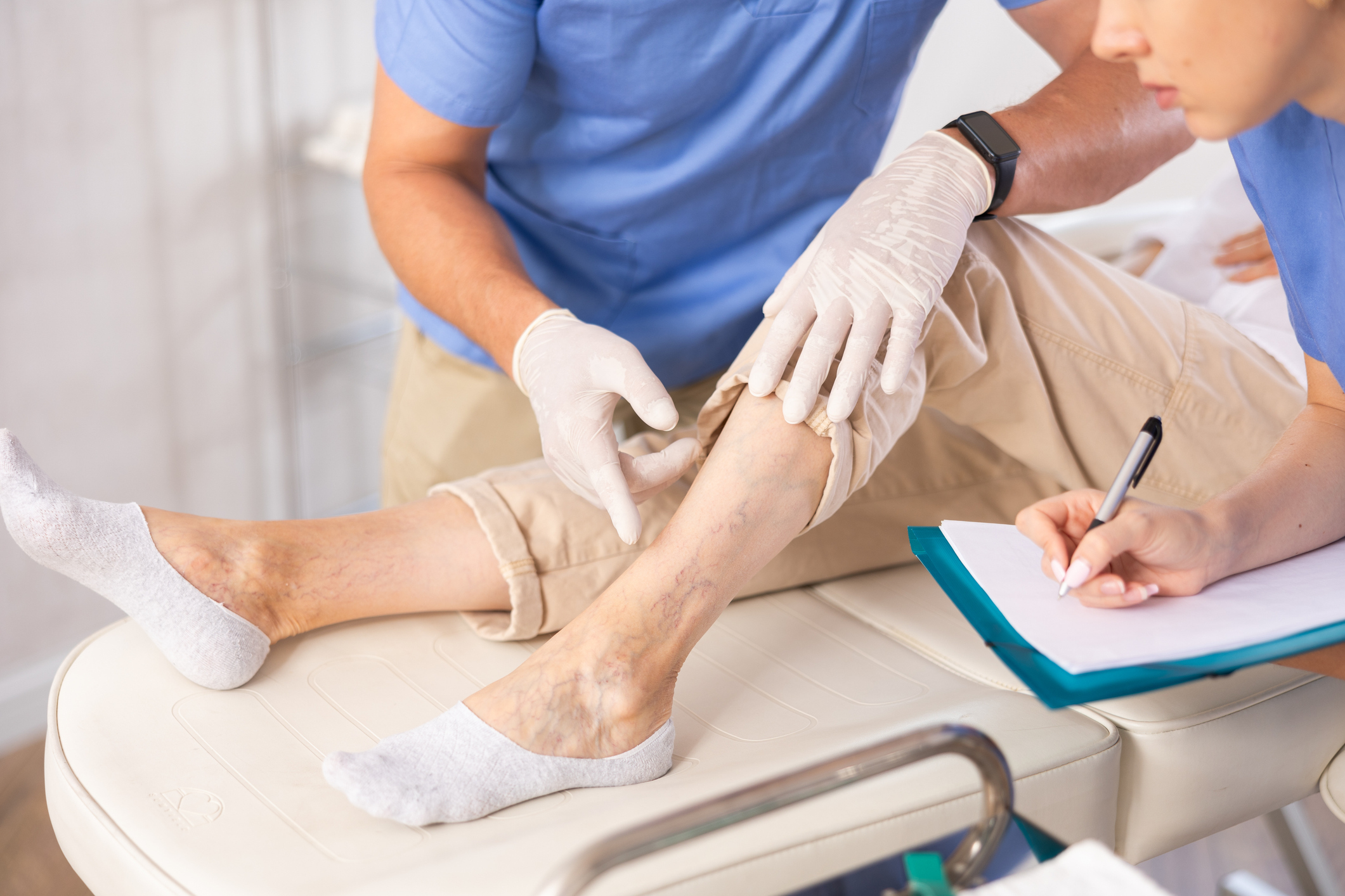 Close-up male doctor exam legs of senior woman against varicose veins, before sclerotherapy treatment. Nurse filling the document.
